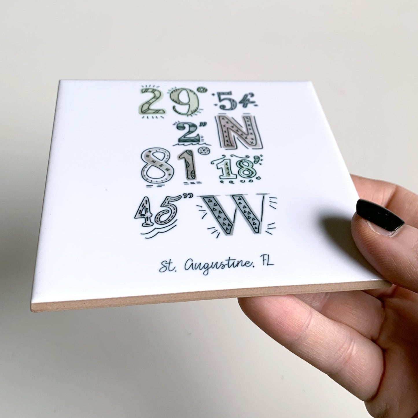 A hand holding a close-up of an illustrated city art coaster, showing the ceramic tile edges