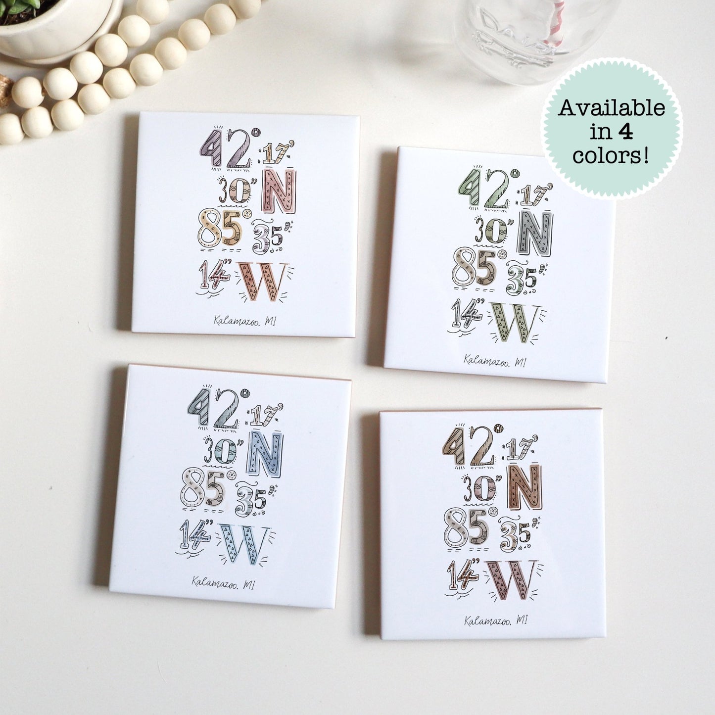 A city drawing of the coordinates of Kalamazoo MI a on a set of ceramic coasters in four colors, sitting on a table