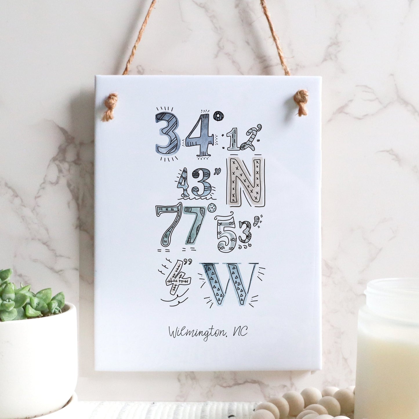 A city drawing of the coordinates of Wilmington NC on a rectangle tile sign, hanging on a wall, in the color coastal