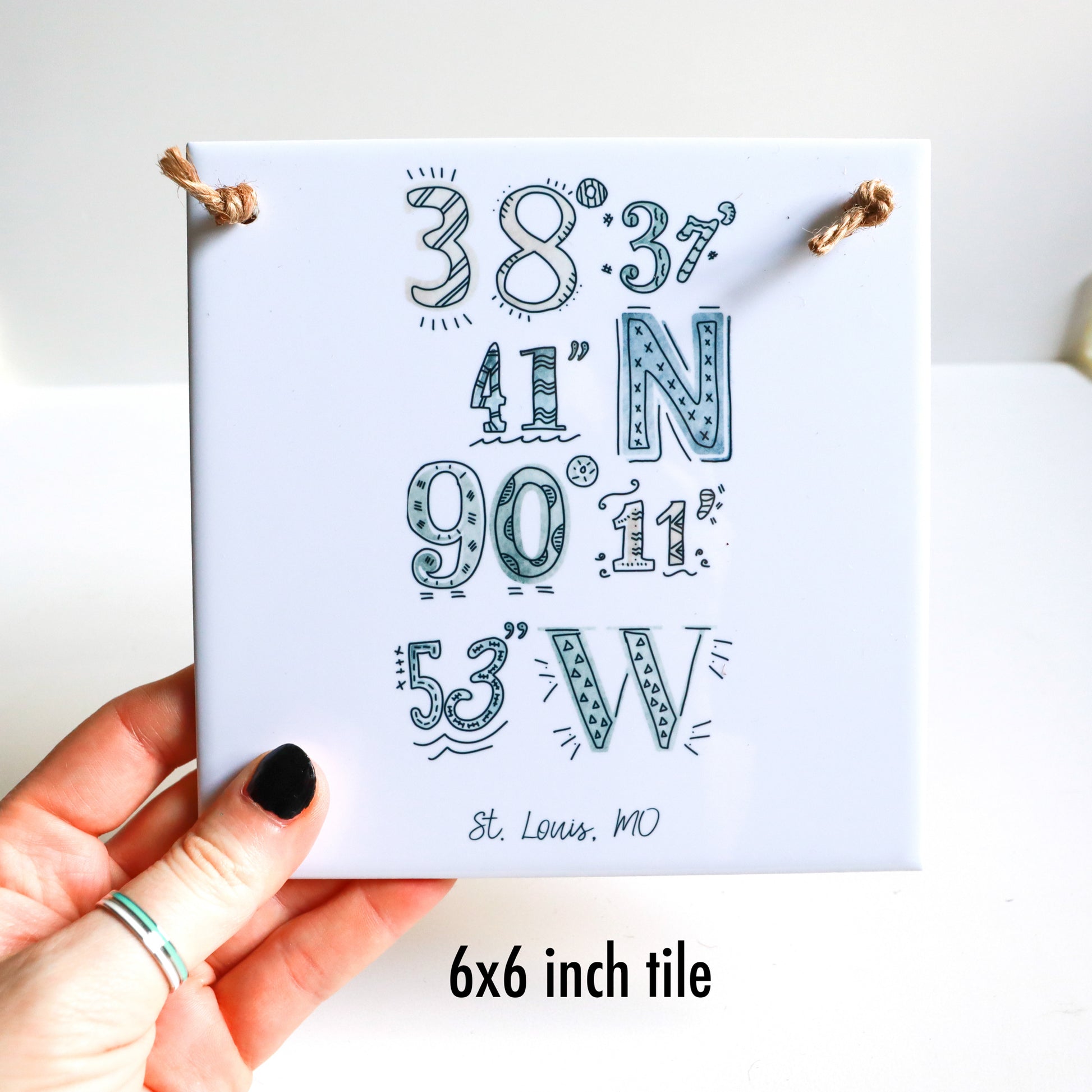 A hand holding the 6x6 in ceramic tile sign, featuring a custom city art coordinates drawing.