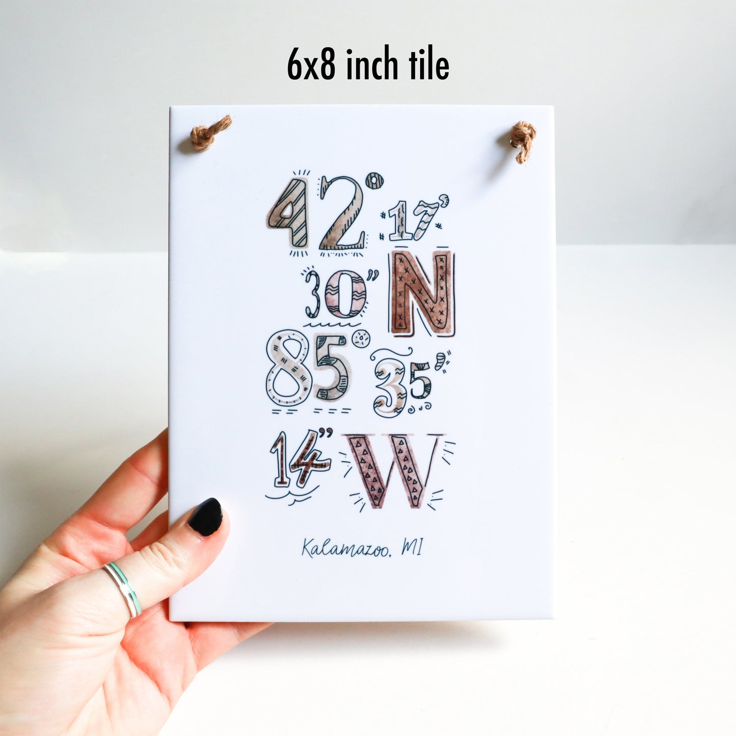 A hand holding the 6x8 in ceramic tile sign, featuring a custom city art coordinates drawing