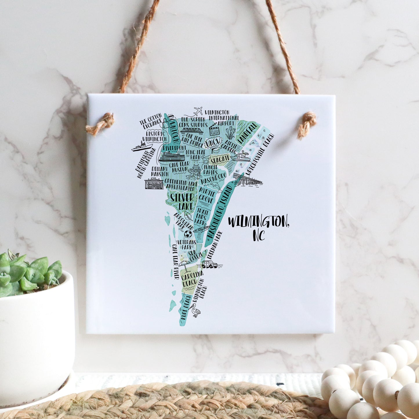 A hand-drawn tourist map of Wilmington North Carolina on a square tile sign hanging on a wall - in the color teal