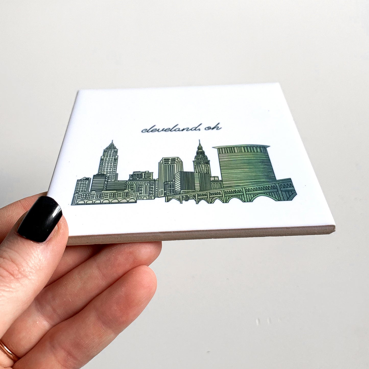 A hand holding a ceramic coaster with a skyline drawing on it, showing the tile edges.