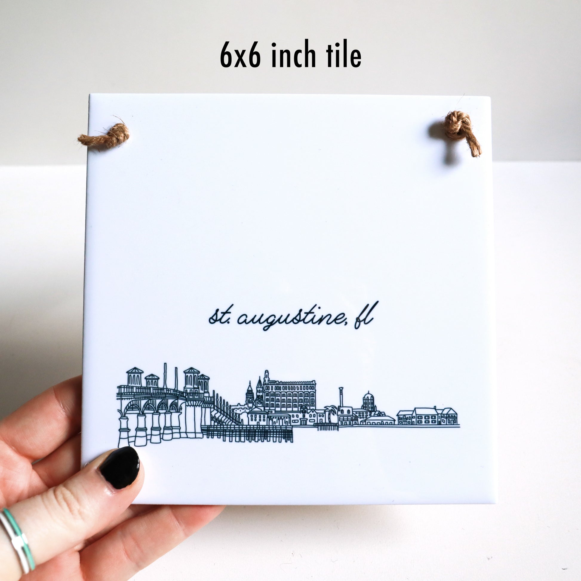 A hand holding a city drawing on a square tile sign, showing the 6x6 inch ceramic tile