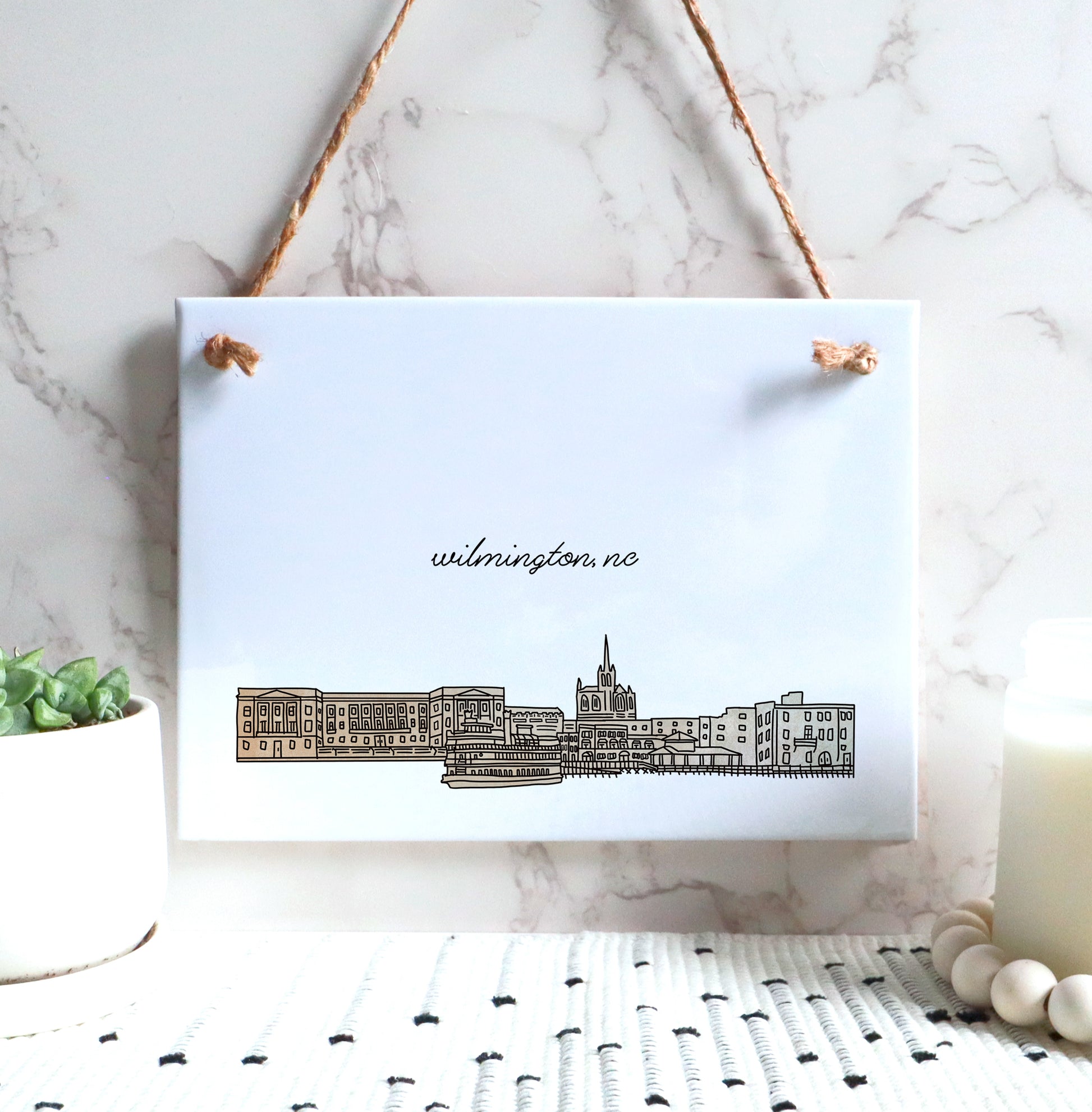 A Wilmington NC souvenir of a drawing of the city skyline on a rectangle tile sign, hanging on a wall - in the color beige