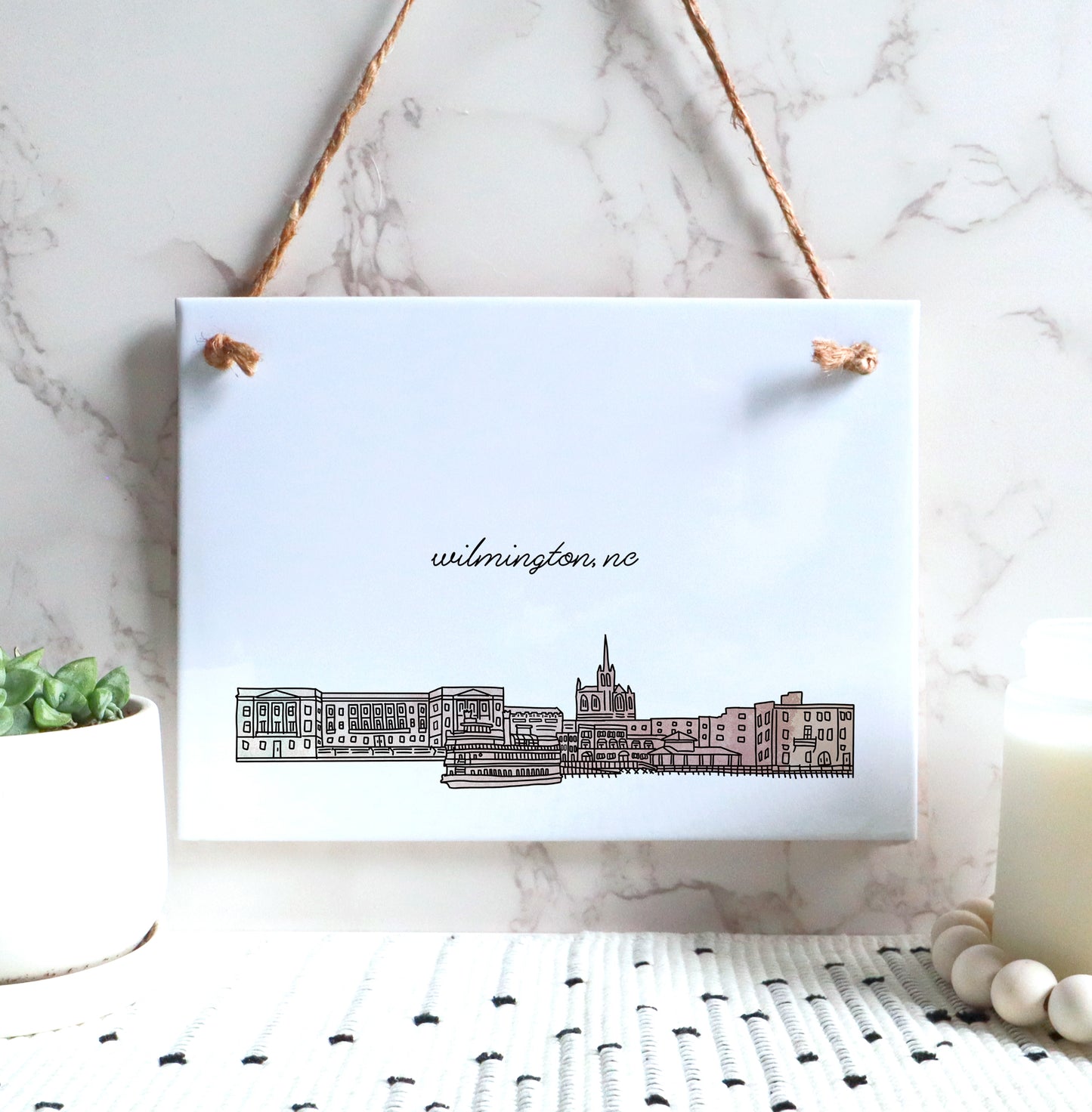 A Wilmington NC souvenir of a drawing of the city skyline on a rectangle tile sign, hanging on a wall - in the color blush