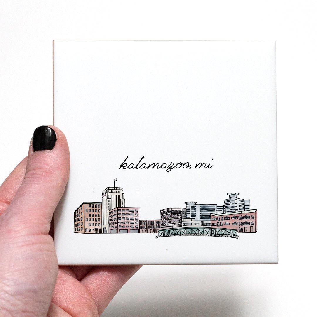 A hand holding a ceramic coaster with a city drawing of Kalamazoo MI - in the color natural