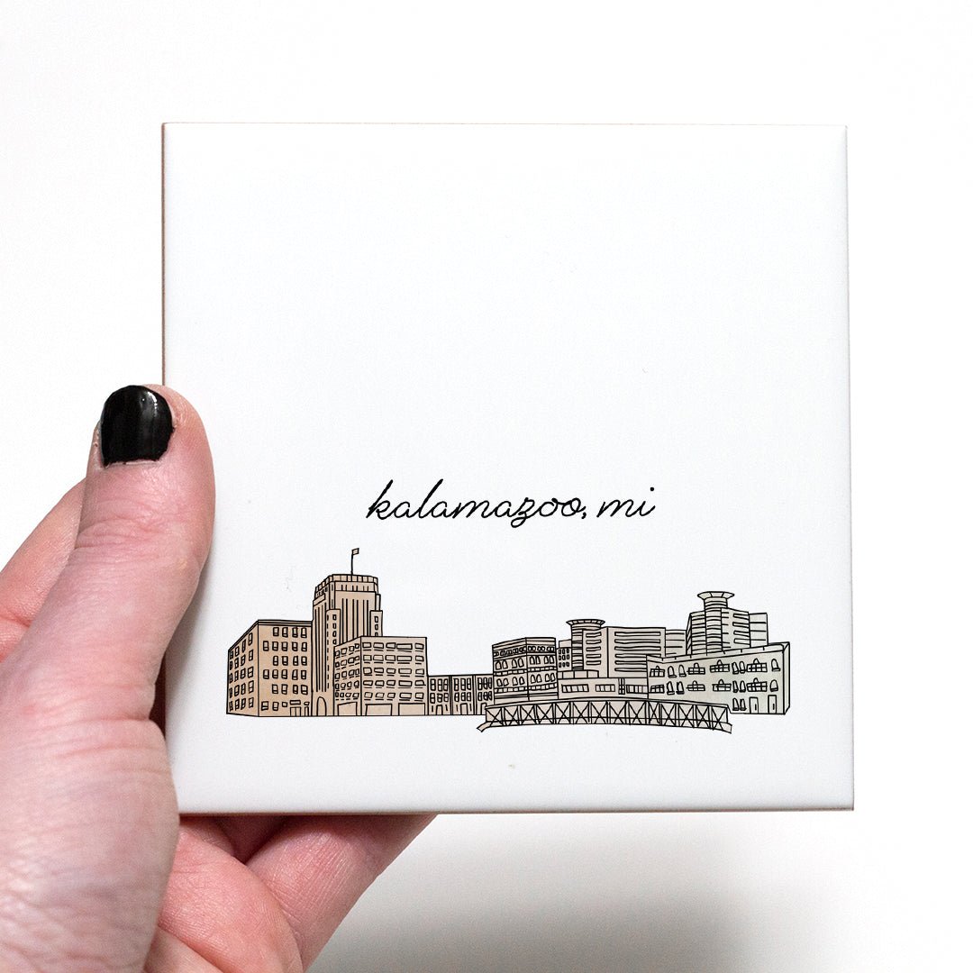 A hand holding a ceramic coaster with a city drawing of Kalamazoo MI - in the color beige