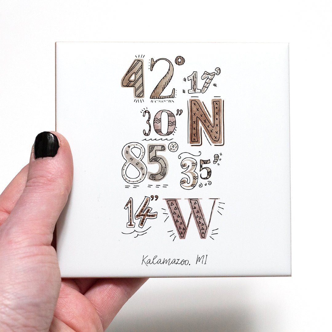 A hand holding a coaster with a city drawing of the coordinates of Kalamazoo Michigan, in the color boho