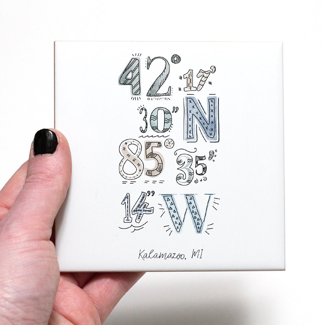 A hand holding a coaster with a city drawing of the coordinates of Kalamazoo Michigan, in the color coastal