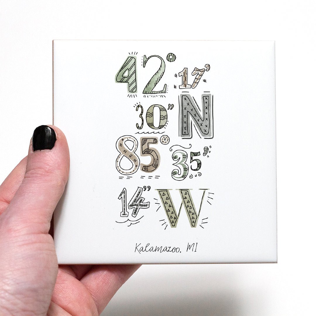A hand holding a coaster with a city drawing of the coordinates of Kalamazoo Michigan, in the color earthy
