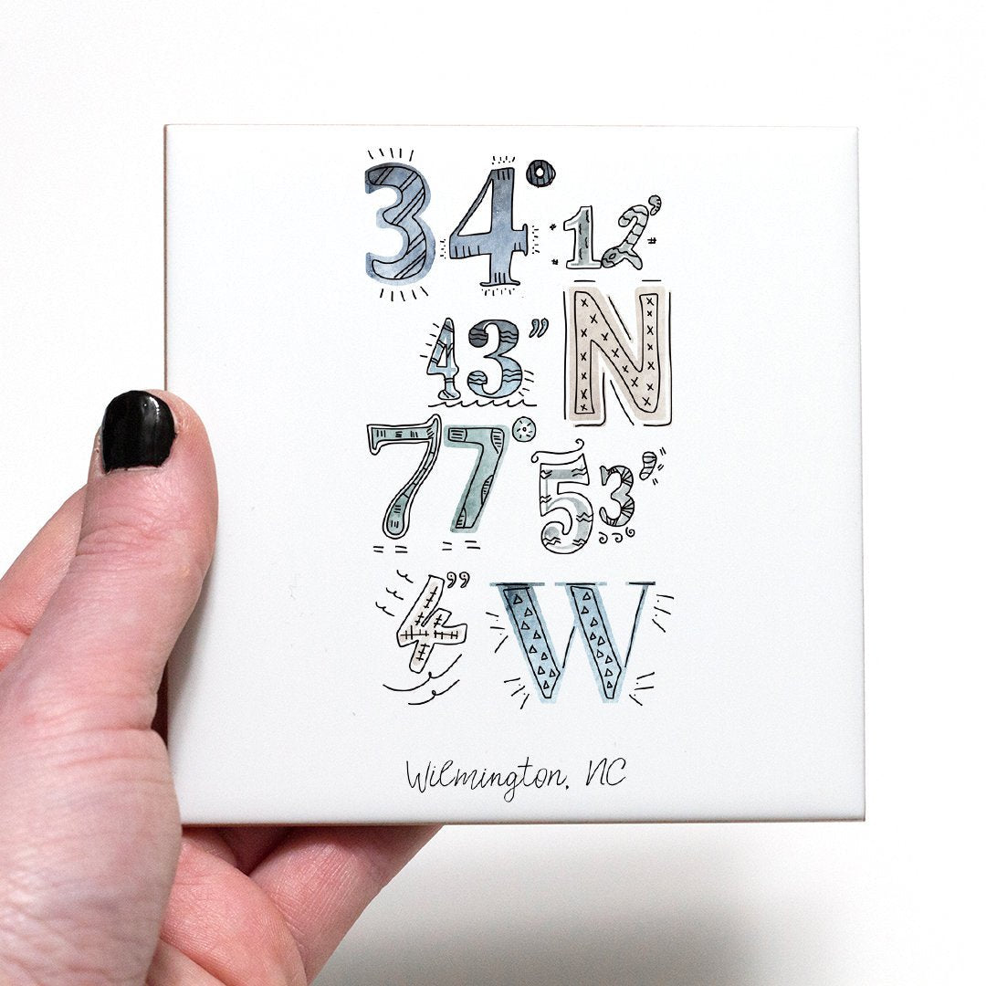 A hand holding a coaster with a city drawing of the coordinates of Wilmington North Carolina, in the color coastal